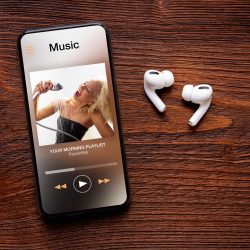 Music player on screen of iPhone and AirPods Pro on wooden surface.